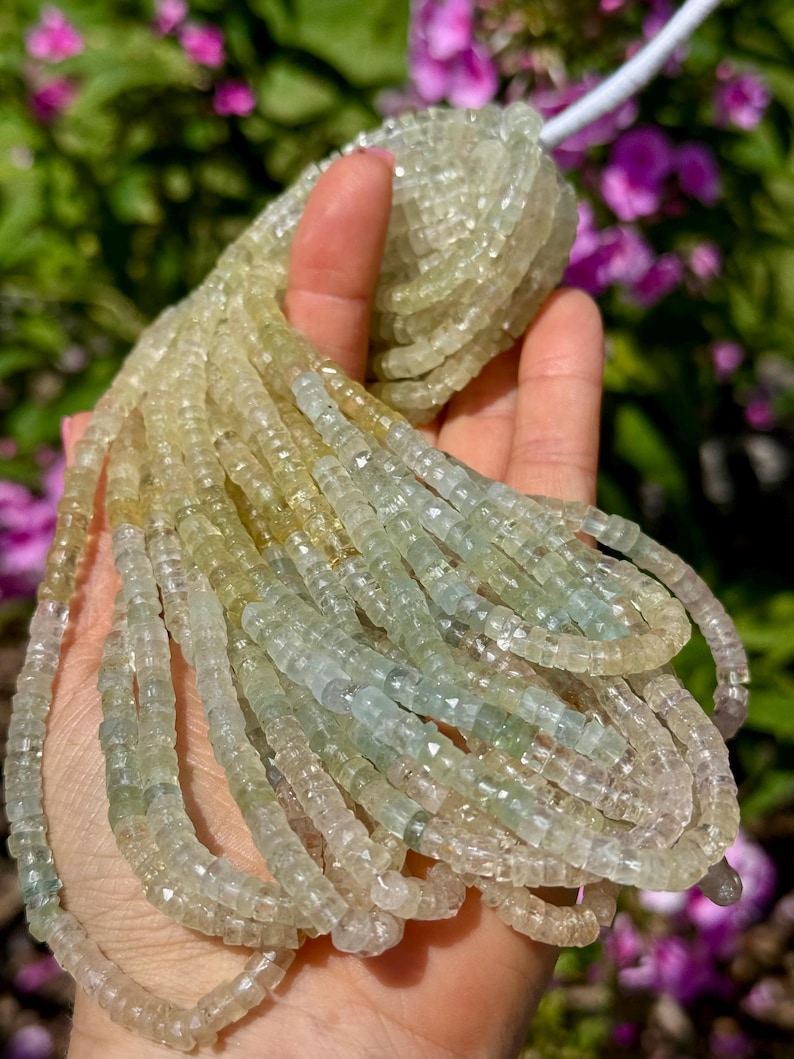 Pu&ograve; includere: Fili di perline di pietre preziose verde chiaro e giallo. Le perline sono piccole, tagliate a forma quadrata e infilate insieme. Le perline sono tenute in una mano, con uno sfondo sfocato di fogliame verde e viola.