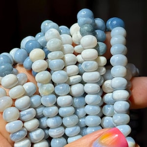 May include: Close-up of several strands of light blue and white gemstone beads. The beads are round and have a smooth, polished finish. The color variations range from pale blue to creamy white, creating a soft, natural aesthetic. The beads are strung together, ready for jewelry making.