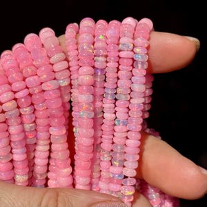 May include: Strands of pink opal beads in varying shades of pink, from light to deep rose. The beads are small, round, and have a subtle iridescent quality. The beads are held in a hand against a black background.