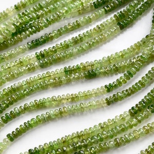 May include: A strand of green gemstone beads, likely peridot, with a faceted, slightly oval shape. The beads are strung on a clear thread and are arranged in a loose, flowing pattern.