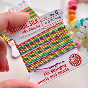 May include: A spool of rainbow colored natural silk thread for stringing pearls and beads. The spool is labeled "NATURAL SILK 2 m No. 6 Ø 0,70 mm 100% Naturseide" and "For stringing pearls and beads".