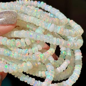 May include: A collection of strands featuring small, round, disc-shaped white opal beads. Each bead displays a translucent quality with iridescent flashes of green, blue, and pink light reflecting across the surface of the polished stone material.