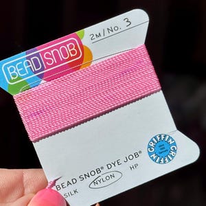 May include: A spool of pink nylon thread labeled "BEAD SNOB" and "DYE JOB". The card also features the Griffin logo and text indicating 2 meters of thread, size 3. The thread is silk and nylon.