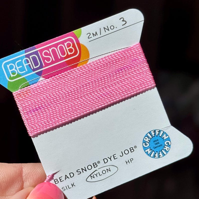 May include: A spool of pink nylon thread labeled "BEAD SNOB" and "DYE JOB". The card also features the Griffin logo and text indicating 2 meters of thread, size 3. The thread is silk and nylon.