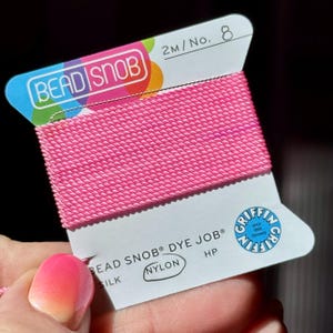 May include: A spool of bright pink nylon thread labeled "BEAD SNOB" and "DYE JOB." The label also includes "SILK" and "NYLON" text. The thread is 2 meters in length and is No. 8. The label has a blue and white design with the Griffin logo.