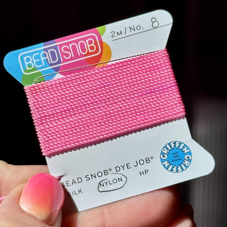 May include: A spool of bright pink nylon thread labeled "BEAD SNOB" and "DYE JOB." The label also includes "SILK" and "NYLON" text. The thread is 2 meters in length and is No. 8. The label has a blue and white design with the Griffin logo.