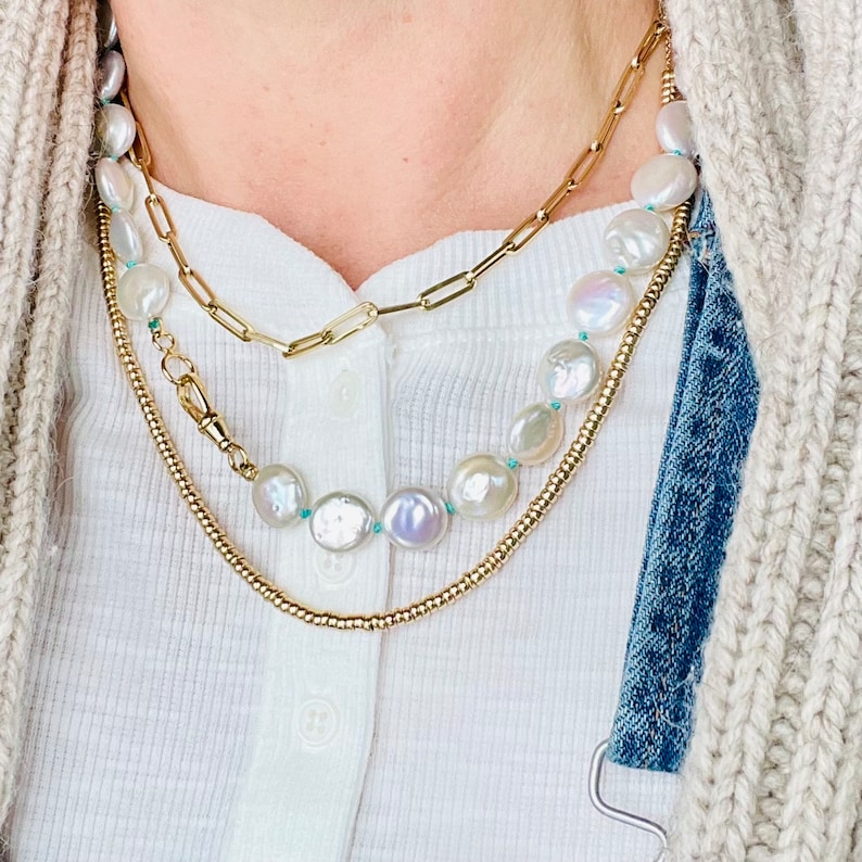 May include: Three gold necklaces layered on a person's neck. The top necklace is a chain link style. The middle necklace is a thin chain with a clasp. The bottom necklace is a thicker chain with a pearl strand. The pearls are white with a turquoise accent.