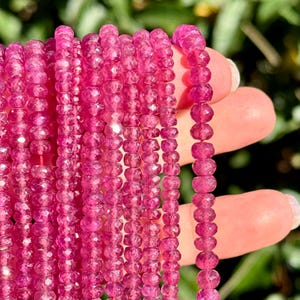 May include: A collection of multiple strands of faceted, vibrant magenta-pink gemstone beads. Each bead features a circular, flat-sided shape with a polished, reflective surface that catches the light, arranged in long, vertical, parallel rows.