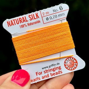 May include: A spool of 2 meters of orange natural silk thread, 0.70 mm in diameter, labeled "NATURAL SILK 100% Naturseide" and "No. 6". The spool is labeled "For stringing pearls and beads" and "www.griffin.de".
