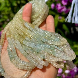 Pu&ograve; includere: Fili di perline di pietre preziose verde chiaro e giallo. Le perline sono piccole, tagliate a forma quadrata e infilate insieme. Le perline sono tenute in una mano, con un filo bianco visibile in alto. Lo sfondo &egrave; sfocato, con accenni di verde e viola.