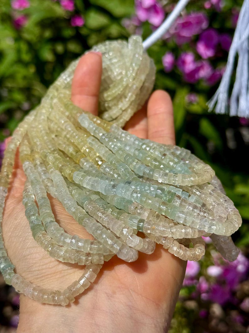 Pu&ograve; includere: Fili di perline di pietre preziose verde chiaro e giallo. Le perline sono piccole, tagliate a forma quadrata e infilate insieme. Le perline sono tenute in una mano, con un filo bianco visibile in alto. Lo sfondo &egrave; sfocato, con accenni di verde e viola.