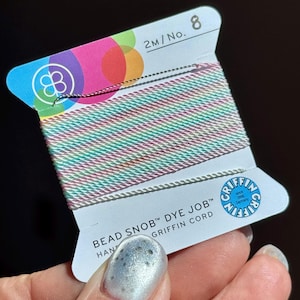 Peut inclure: Une bobine de fil Griffin Bead Snob Dye Job aux couleurs pastel d&eacute;grad&eacute;es, dont rose, vert et blanc. L'&eacute;tiquette indique "2M/No. 8" et comprend le logo Griffin. Le fil est tenu dans une main.
