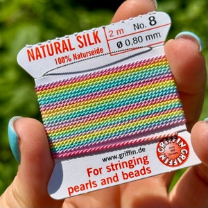 May include: A spool of rainbow colored natural silk thread for stringing pearls and beads. The label reads "NATURAL SILK 100% Naturseide 2m No. 8 Ø 0,80 mm www.griffin.de For stringing pearls and beads GRIFFIN since 1866 Germany".