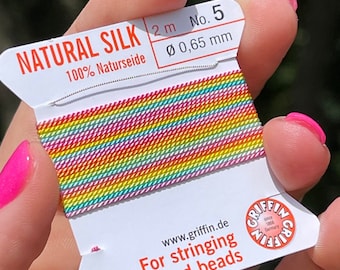 May include: A spool of rainbow coloured natural silk thread for stringing pearls and beads. The spool is labelled "NATURAL SILK 2m No. 5 100% Naturseide Ø 0,65 mm" and "www.griffin.de For stringing pearls and beads".