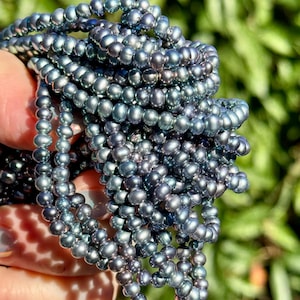 May include: Close-up of strands of dark grey freshwater pearls. The pearls are small and round, with a subtle lustre. The strands are held together, creating a dense, textured appearance. The background is a soft green.