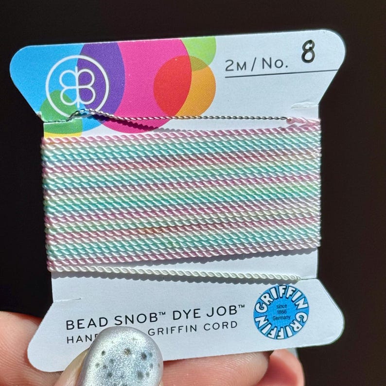 Peut inclure: Une bobine de fil Griffin Bead Snob Dye Job aux couleurs pastel. Le fil est un m&eacute;lange de rose, de vert et de blanc. L'&eacute;tiquette affiche le logo Griffin et le texte "2M/No. 8". Le fil est tenu par une main.