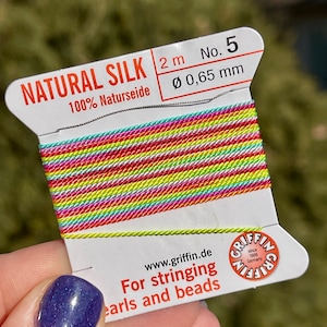 May include: A spool of 2 meters of natural silk thread, 100% Naturseide, with a diameter of 0.65 mm. The thread is rainbow colored and is labeled "NATURAL SILK 2 m No. 5 Ø 0,65 mm 100% Naturseide" and "For stringing pearls and beads".