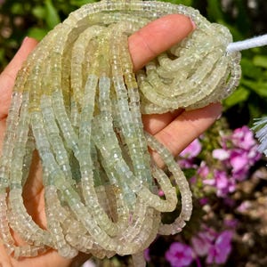 Pu&ograve; includere: Fili di perline di pietre preziose verde chiaro e giallo. Le perline sono piccole, tagliate a forma quadrata e infilate insieme. Le perline sono tenute in una mano, con un filo bianco visibile in alto. Lo sfondo &egrave; sfocato con fogliame verde e rosa.