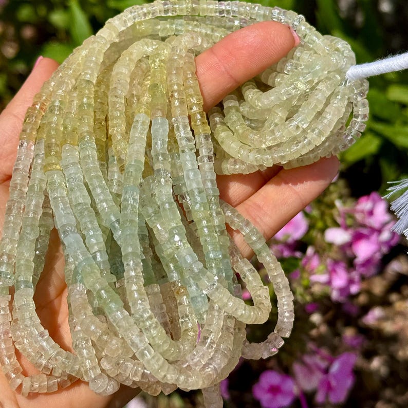 Pu&ograve; includere: Fili di perline di pietre preziose verde chiaro e giallo. Le perline sono piccole, tagliate a forma quadrata e infilate insieme. Le perline sono tenute in una mano, con un filo bianco visibile in alto. Lo sfondo &egrave; sfocato con fogliame verde e rosa.