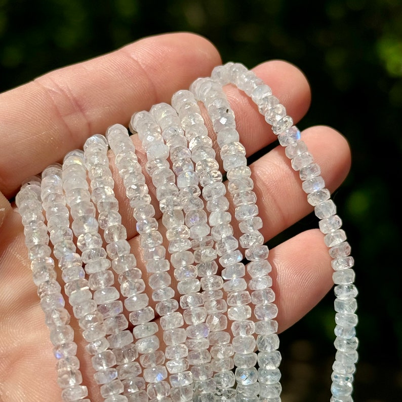 May include: A strand of white moonstone beads, faceted and iridescent, with a rainbow sheen. The beads are small and round, and the strand is long enough to be used for jewelry making.