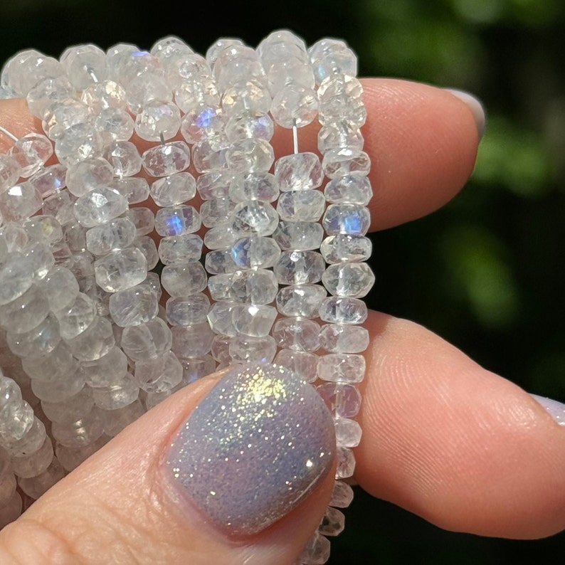 May include: A strand of white moonstone beads with iridescent shimmer. The beads are faceted and have a slightly irregular shape. The beads are strung on a clear thread.