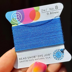 May include: A spool of vibrant blue Griffin Bead Snob dye job silk thread. The label indicates 2m/No. 8, 0.80mm. The packaging features a colorful design and the Griffin logo, showcasing the brand's heritage since 1866 in Germany. The thread is 100% natural silk.