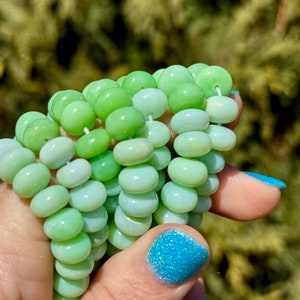 Op de afbeelding: Een streng groene opaal kralen, met een kleurverloop van lichtgroen naar wit. De kralen zijn rond en glad, en ze zijn geregen op een wit draad.