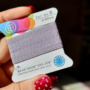 May include: A spool of light purple Griffin Bead Snob Dye Job thread. The label reads "2M/ No. 8 0.80mm" and "Griffin 100% Natural Silk". The label has a colorful design and the Griffin logo. The thread is held in a hand with red nail polish.