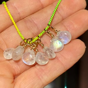 Op de afbeelding: Een ketting met meerdere iriserende maansteen hangers. De druppelvormige hangers zijn opgehangen aan goudkleurige ringen aan een neon groen koord. De maanstenen tonen een reeks kleuren, waaronder wit, blauw en geel.