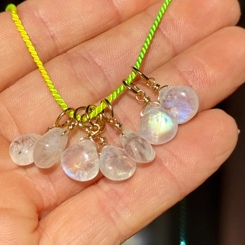Op de afbeelding: Een ketting met meerdere iriserende maansteen hangers. De druppelvormige hangers zijn opgehangen aan goudkleurige ringen aan een neon groen koord. De maanstenen tonen een reeks kleuren, waaronder wit, blauw en geel.