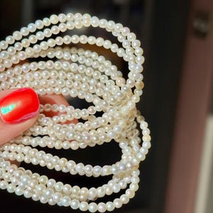 May include: Close-up of multiple strands of small, round, white pearls. The pearls are tightly strung, creating a delicate look. The pearls are held by a hand with red nail polish.
