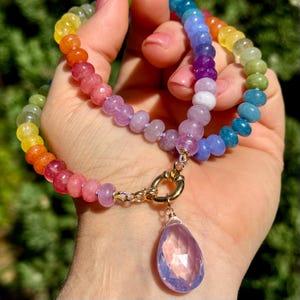 May include: A rainbow gemstone necklace with a large pink teardrop pendant. The necklace is made of round beads in various colors, including yellow, orange, red, pink, purple, blue, and green.