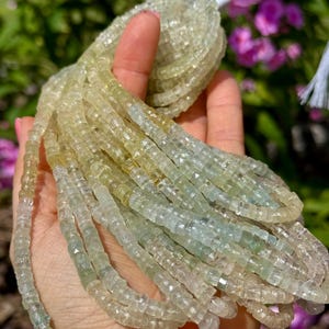 Pu&ograve; includere: Fili di perline di pietre preziose verde chiaro e giallo. Le perline sono piccole, tagliate a forma quadrata e infilate insieme. Le perline sono tenute in una mano, con uno sfondo sfocato di fogliame verde e fiori rosa.