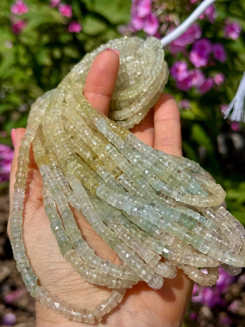 Pu&ograve; includere: Fili di perline di pietre preziose verde chiaro e giallo. Le perline sono piccole, tagliate a forma quadrata e infilate insieme. Le perline sono tenute in una mano, con uno sfondo sfocato di fogliame verde e fiori rosa.