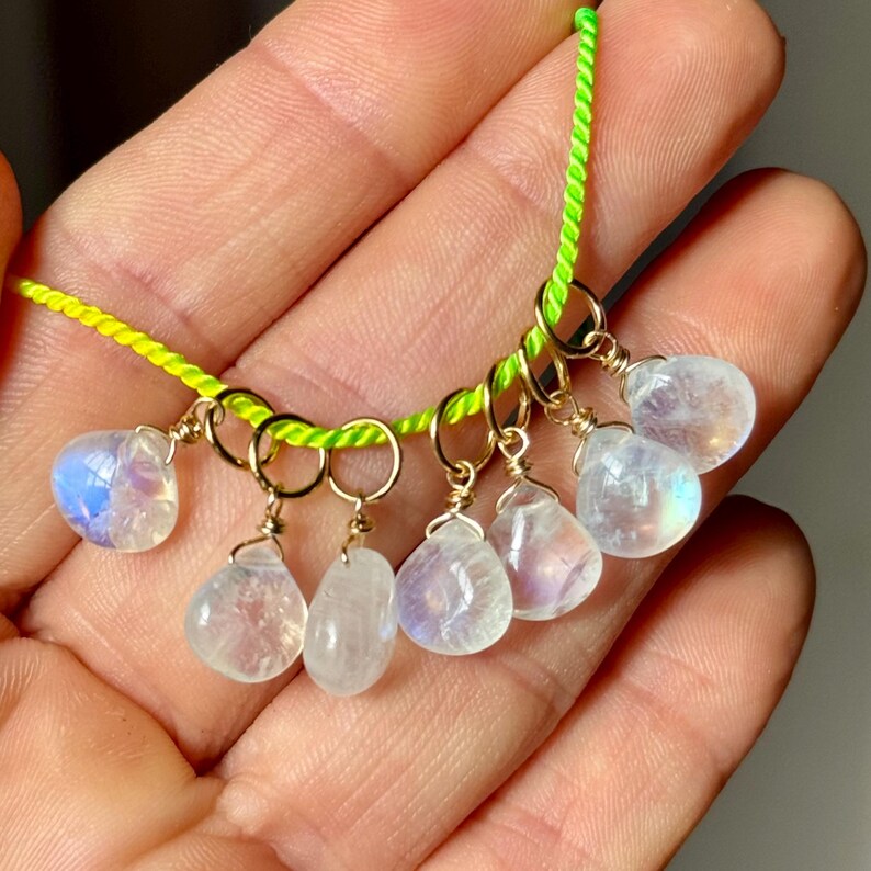 Op de afbeelding: Een ketting met zeven druppelvormige maansteen hangers. Elke steen is met gouddraad omwikkeld en hangt aan een neon groen koord. De maanstenen vertonen een subtiele iriserende glans en de gouden draad voegt elegantie toe.