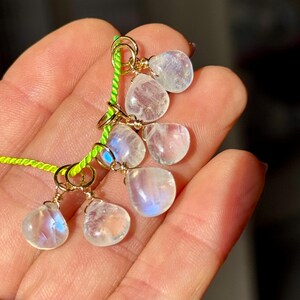 Op de afbeelding: Een cluster van druppelvormige maansteenhangers, elk met een gouden draadwikkeling, is geregen aan een felgroen koord. De maanstenen vertonen een zachte, iriserende gloed. De sieraden worden in een hand gehouden, met de achtergrond onscherp.