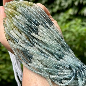 May include: Strands of faceted gemstone beads in shades of blue, green, and gray. The beads are small and strung together, creating a flowing, textured appearance. The background is a soft green, suggesting an outdoor setting.