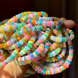 Puede incluir: Una colección de collares de cuentas coloridas en tonos rosa, naranja, amarillo, verde y azul. Las cuentas son pequeñas y redondas, ensartadas para formar múltiples hebras. Los collares se sostienen en una mano, con la luz reflejándose en las cuentas.