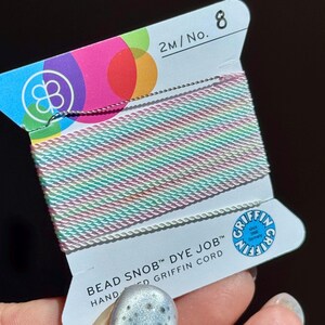 Peut inclure: Une bobine de cordon de perles Griffin aux couleurs pastel, rose, vert et blanc. L'&eacute;tiquette indique "BEAD SNOB DYE JOB" et "HAND GRIFFIN CORD". Le cordon mesure 2 m&egrave;tres, No. 8.
