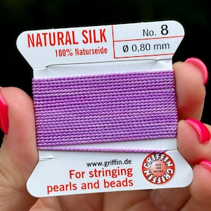 May include: A spool of purple natural silk thread for stringing pearls and beads. The spool is labeled "NATURAL SILK 100% Naturseide No. 8 Ø 0,80 mm" and "www.griffin.de For stringing pearls and beads".