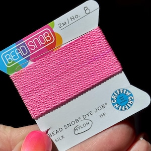 Puede incluir: Un carrete de hilo de nailon rosa brillante para abalorios, etiquetado como "BEAD SNOB DYE JOB". La etiqueta también indica "SILK" y "NYLON" con el logotipo de Griffin. El hilo mide 2 metros de largo, No. 8.