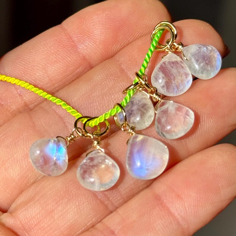 Op de afbeelding: Een verzameling druppelvormige maansteen kralen met een gouden draadwikkeling, geregen op een neon groen koord. De maanstenen vertonen een subtiele blauwe iriserende glans. De sieraden worden gepresenteerd tegen een warme, neutrale achtergrond.