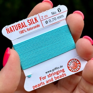 Puede incluir: Un carrete de hilo de seda natural turquesa, etiquetado como "NATURAL SILK" y "No. 6". El embalaje indica una longitud de 2 metros y un diámetro de 0,70 mm. También incluye el texto "For stringing pearls and beads".