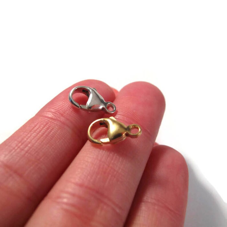 May include: Two lobster claw clasps, one silver and one gold, are shown on a finger.