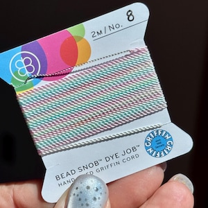 Peut inclure: Une bobine de cordon Griffin Bead Snob Dye Job, avec un m&eacute;lange de fils blancs, roses et turquoise. L'&eacute;tiquette indique "2M/No. 8" et le logo Griffin. Le cordon est teint &agrave; la main en Allemagne.
