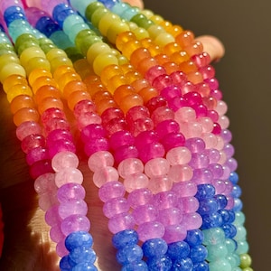 Puede incluir: Primer plano de hilos de cuentas coloridas en un arcoíris de tonos. Las cuentas son redondas y parecen estar hechas de un material translúcido, con colores que van del azul al morado, rosa, naranja, amarillo y verde. Las cuentas están ensartadas.