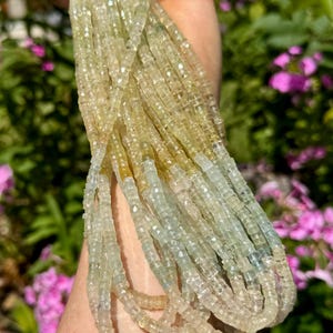 Pu&ograve; includere: Una collezione di fili di perline di pietre preziose giallo chiaro e blu pallido. Le perline sono piccole, tagliate a forma quadrata e infilate insieme. I fili sono tenuti in una mano, con uno sfondo sfocato di fogliame verde e fiori rosa.