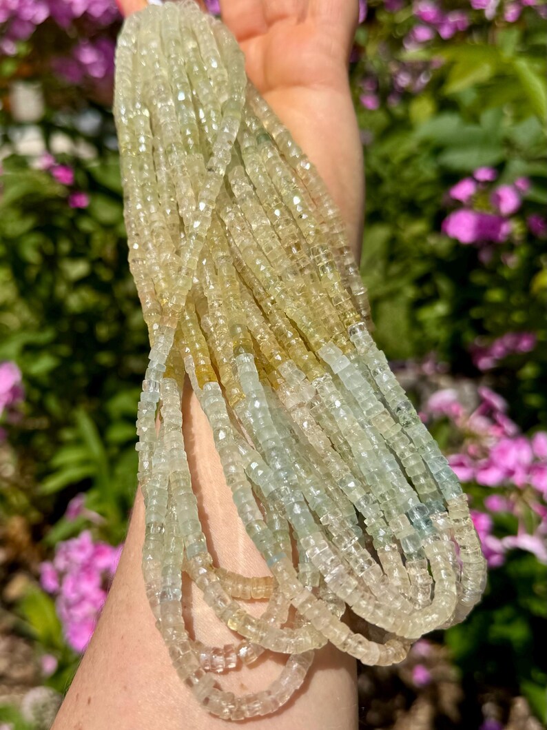 Pu&ograve; includere: Una collezione di fili di perline di pietre preziose giallo chiaro e blu pallido. Le perline sono piccole, tagliate a forma quadrata e infilate insieme. I fili sono tenuti in una mano, con uno sfondo sfocato di fogliame verde e fiori rosa.