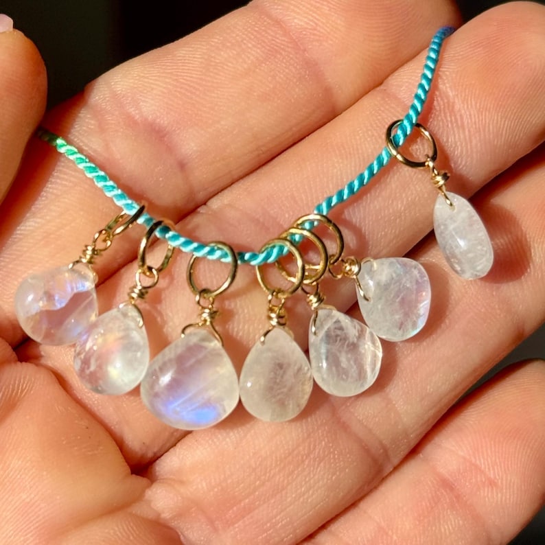 Op de afbeelding: Een delicate ketting met druppelvormige maansteenhangers. De hangers zijn bevestigd aan goudkleurige ringen op een turquoise koord. De maanstenen hebben een doorschijnende, iriserende kwaliteit.