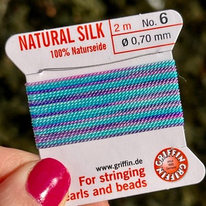 Puede incluir: Una bobina de hilo de seda natural 100% en un esquema de colores azul y morado. El hilo mide 2 metros de largo y 0,70 mm de diámetro. La etiqueta dice "NATURAL SILK 2 m No. 6 100% Naturseide Ø 0,70 mm" y "www.griffin.de Para enhebrar perlas y cuentas".